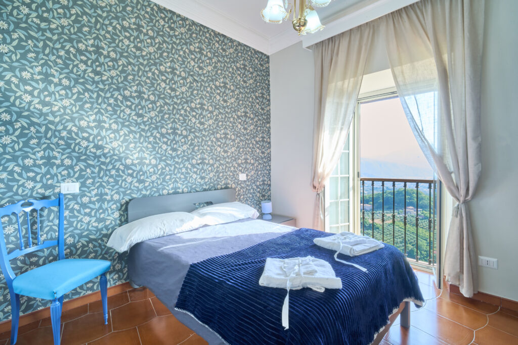 Camera con letto matrimoniale, carta da parati verde a fiori e portafinestra sul balcone