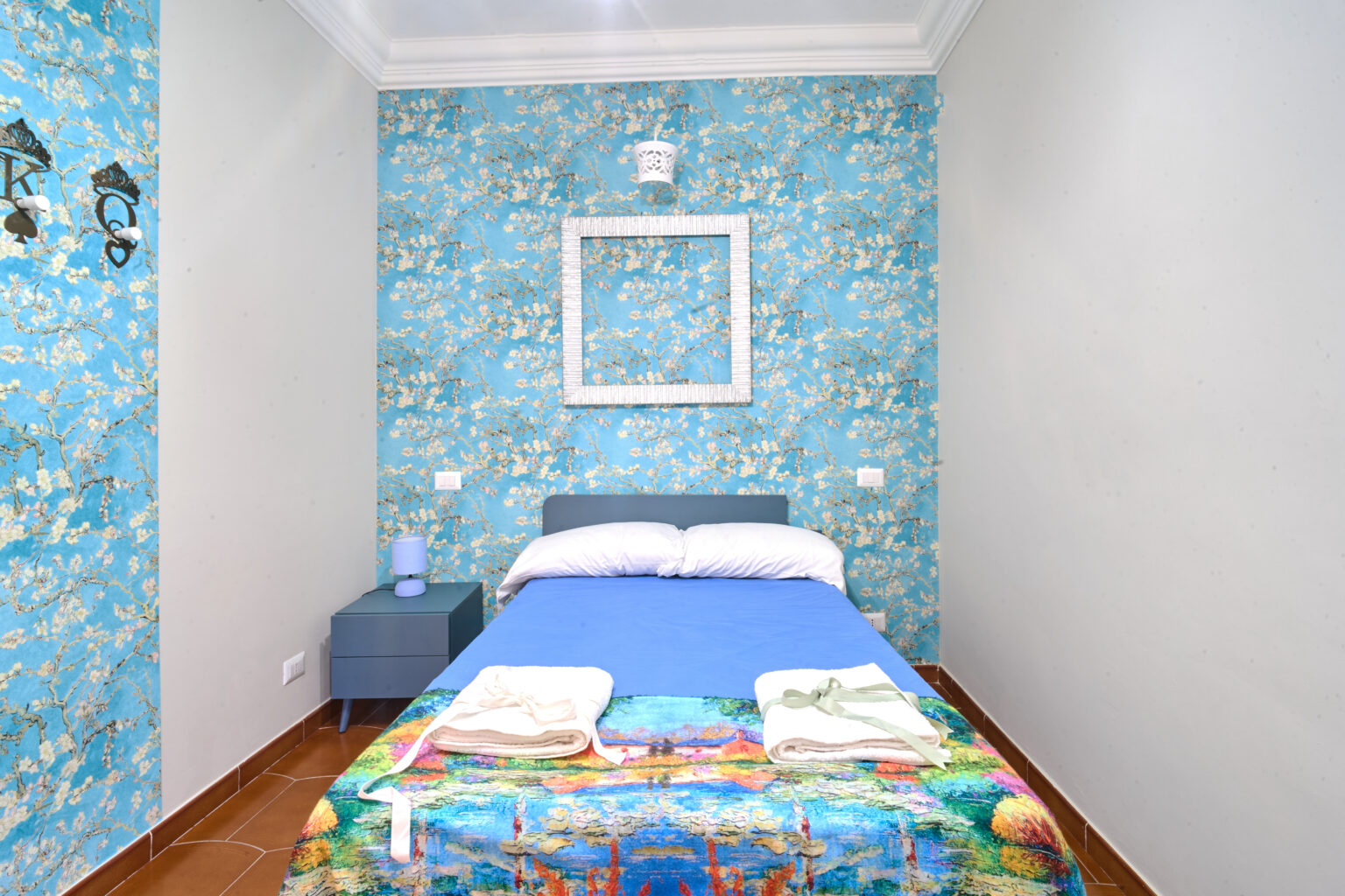 Camera matrimoniale con carta da parati azzurra a fiori e letto con copriletto colorato
